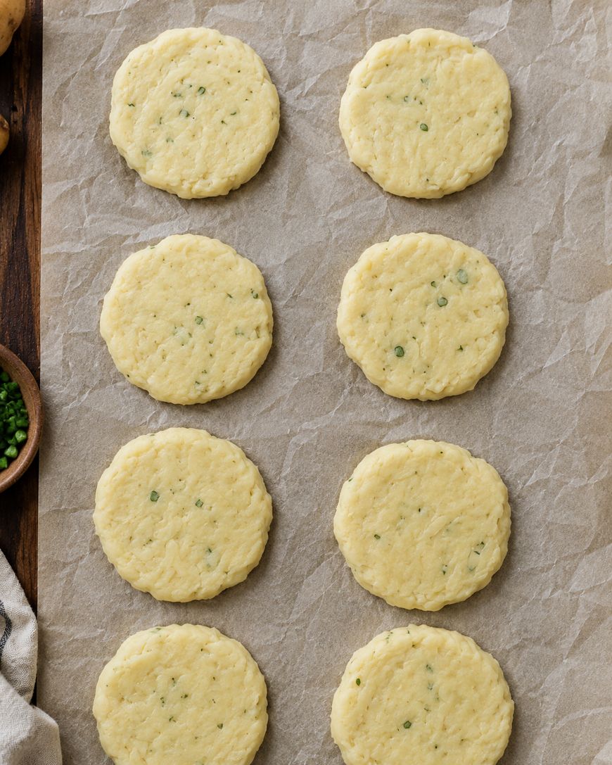 Eight round, unbaked biscuit dough discs with green herb flecks are arranged in two vertical rows on a sheet of parchment paper.