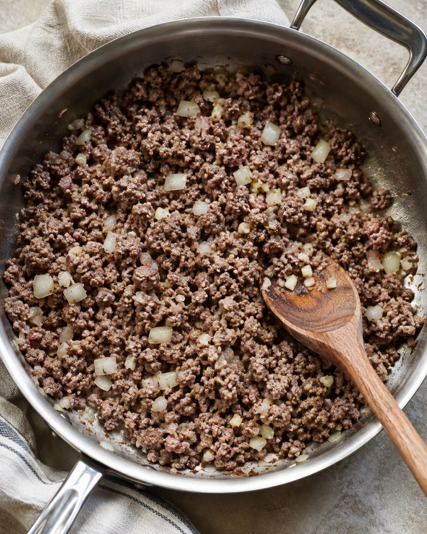 Ground beef and chopped onions being cooked in a stainless steel skillet, stirred with a wooden spoon, with a towel and striped cloth partially visible nearby.