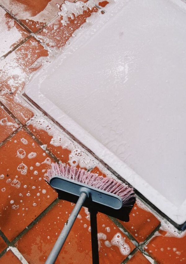 A broom with pink bristles cleaning soapy water from red tiled floor near a white mat.