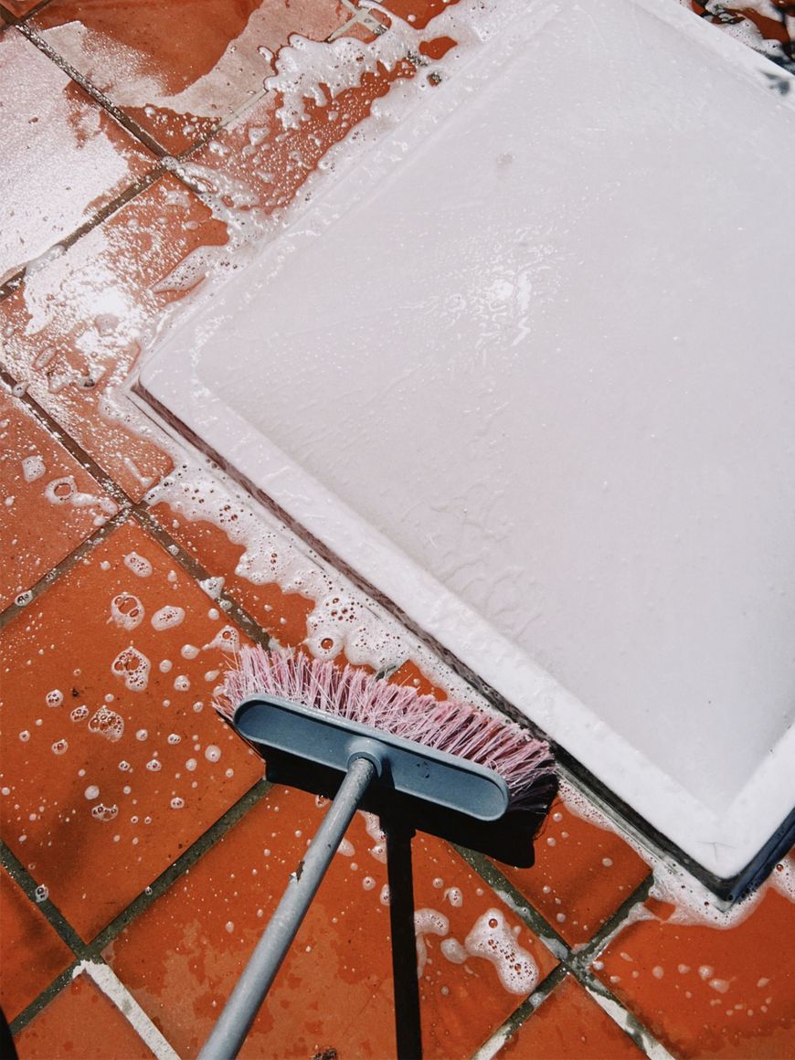 A pink-bristled brush lies on soapy, wet red tiles next to a white rectangular mat, indicating cleaning in progress.