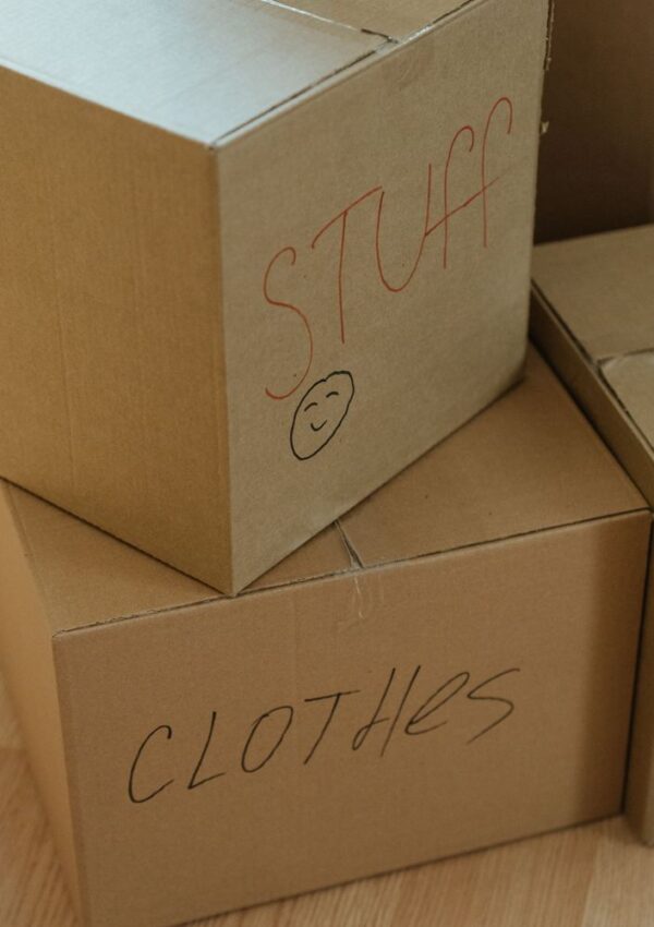Three cardboard boxes stacked. One is labeled STUFF with a smiley face drawn underneath, and another is labeled CLOTHES. The boxes are sitting on a wooden floor.