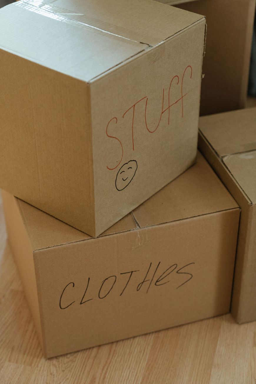 Three cardboard moving boxes are stacked on a wooden floor. One box is labeled STUFF with a smiley face, and another is labeled CLOTHES. Both labels are handwritten in marker.