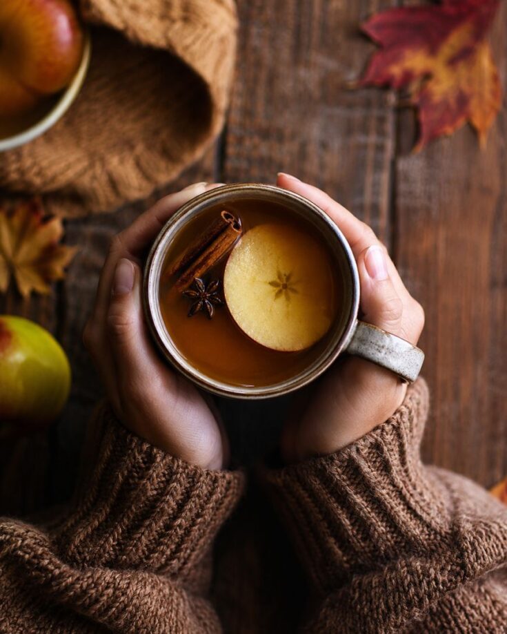Hands in a brown sweater hold a mug of spiced apple cider topped with a cinnamon stick, star anise, and a round apple slice. Autumn leaves and apples are scattered on a rustic wooden table.