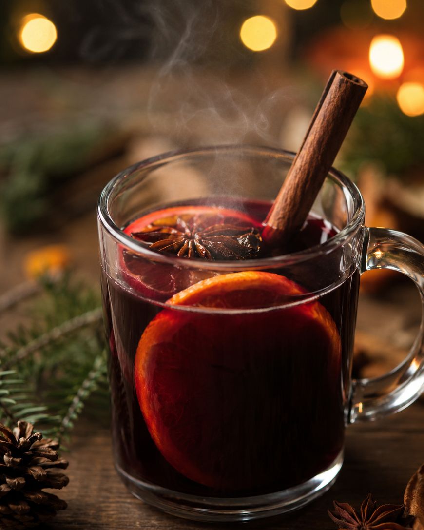 A clear glass mug of steaming mulled wine garnished with a cinnamon stick, star anise, and orange slice, set on a wooden table with pinecones, fir branches, and warm holiday lights in the background.