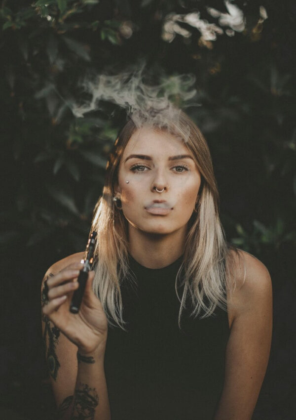 A woman with blonde hair exhales vapor while holding a vape pen, standing outdoors with dark green leaves in the background.