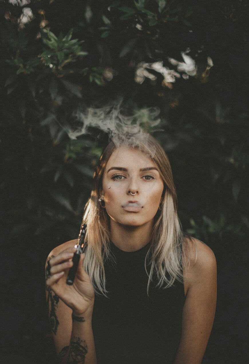 A woman with blonde hair holds a vape pen and exhales vapor, surrounded by dark green foliage. She wears a sleeveless black top and has tattoos on her left arm.