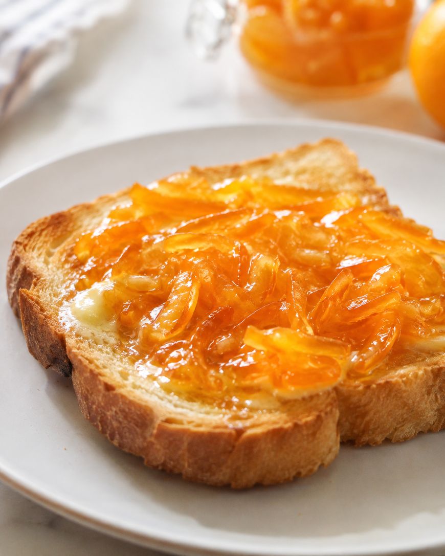 A slice of toasted bread topped with glossy orange marmalade sits on a white plate. A jar of marmalade and part of an orange are blurred in the background.