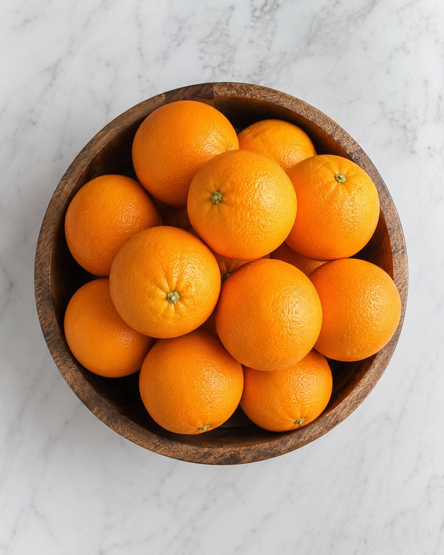 A wooden bowl filled with whole, bright oranges sits on a white marble surface.