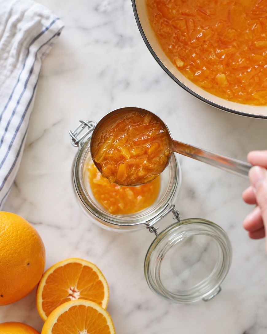 A hand pours orange marmalade from a ladle into a glass jar. Sliced oranges and a pot of marmalade are on a marble countertop, with a striped towel nearby.