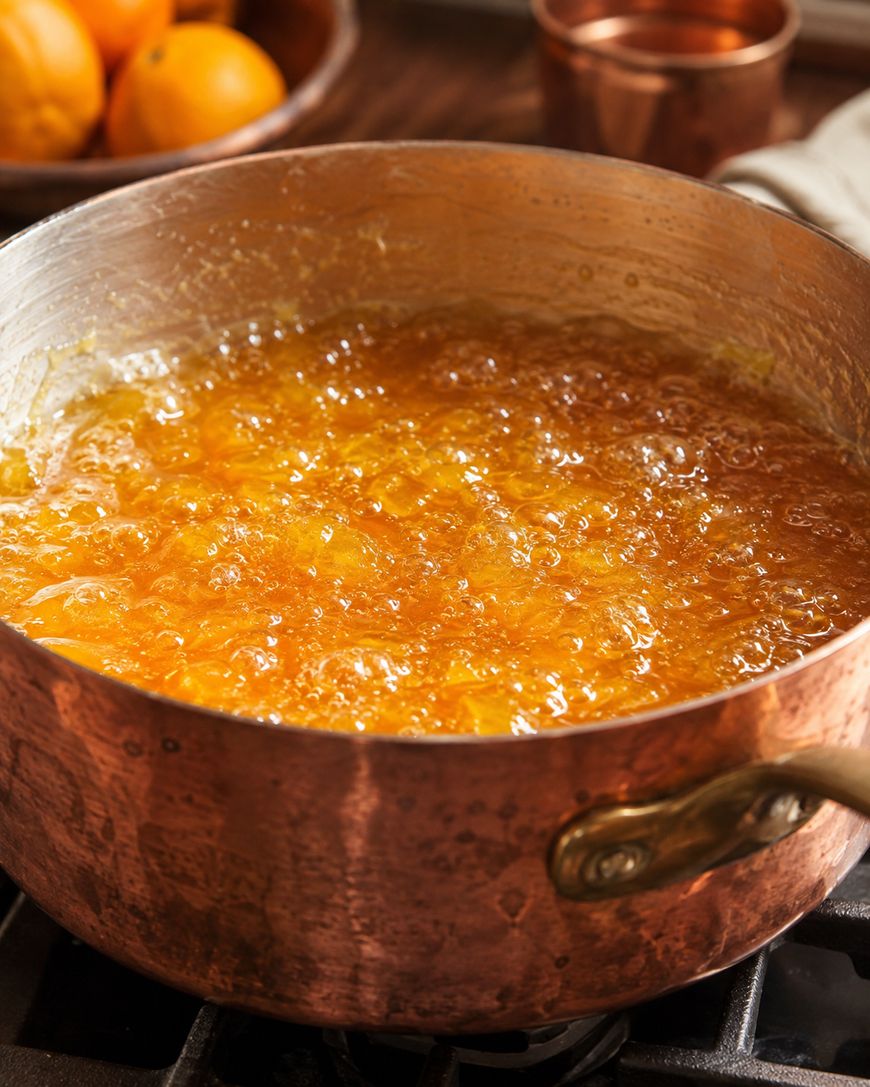 A copper pot filled with bubbling orange marmalade cooks on a stovetop, with oranges and a copper cup blurred in the background.
