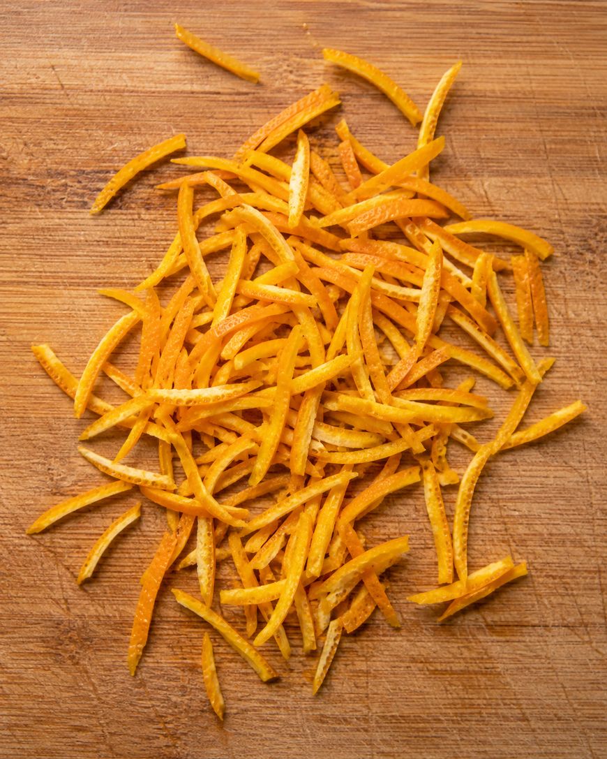 Thin strips of orange zest are scattered on a wooden cutting board, creating a textured and colorful pile.