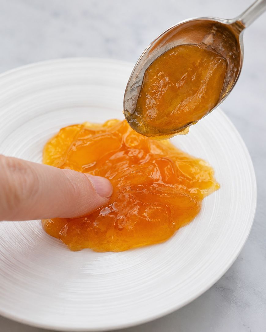 A spoon spreads orange marmalade onto a white plate while a finger gently presses the jelly, showing its texture.