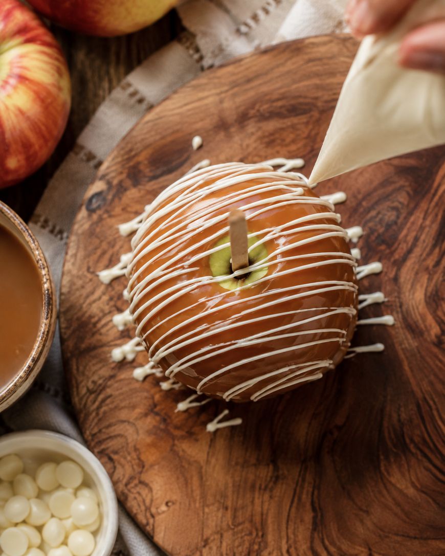 A hand drizzles white chocolate over a caramel apple on a wooden board, surrounded by apples, caramel, and white chocolate chips.