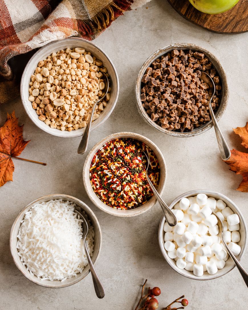 Five small bowls with spoons hold chopped peanuts, chocolate pieces, shredded coconut, mini marshmallows, and fall-colored sprinkles on a light surface with autumn decorations nearby.