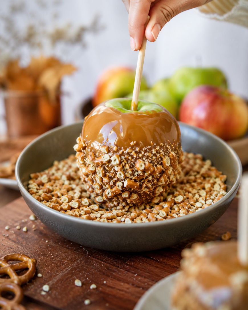 A hand dips a green caramel apple into a bowl of crushed nuts, with apples and pretzels in the background.