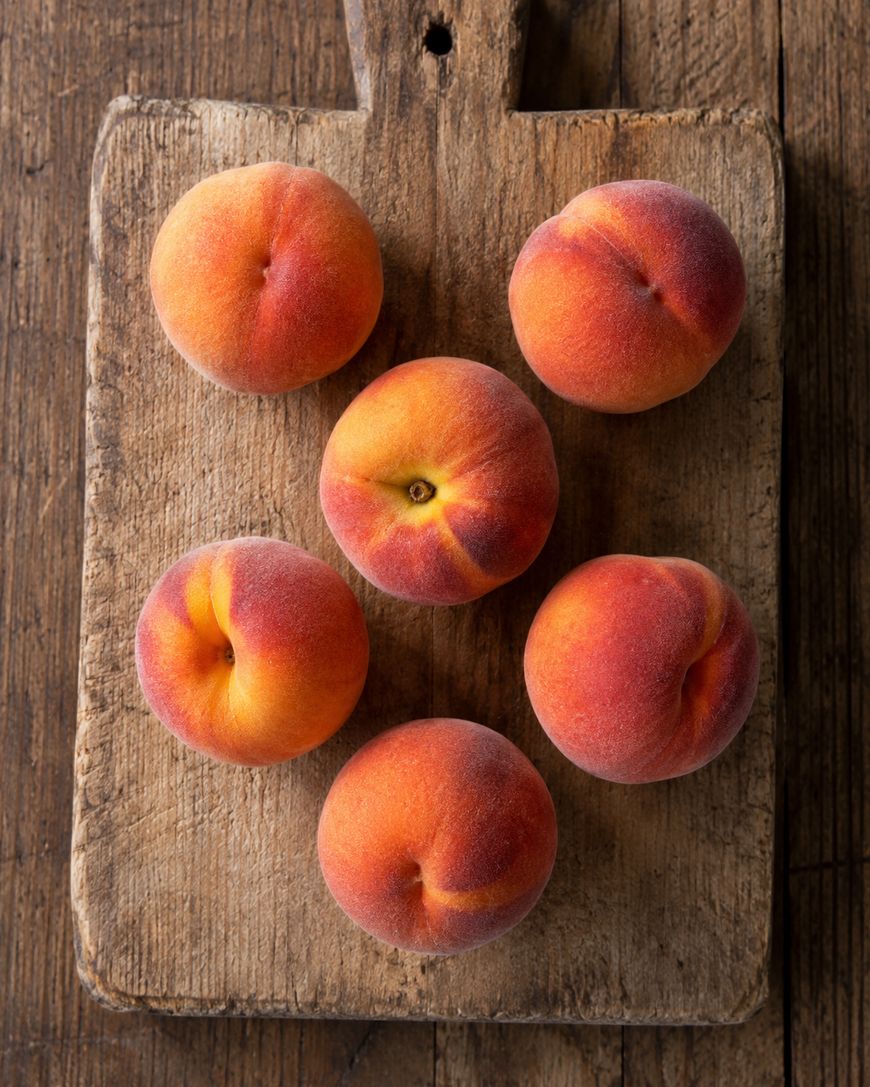 Six ripe peaches are arranged in a circle on a rustic wooden cutting board, which sits on an aged wooden table. The peaches have a fuzzy texture and display shades of orange, red, and yellow.