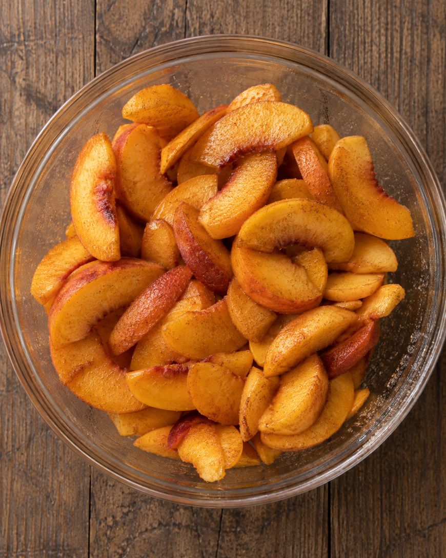 A glass bowl filled with sliced peaches coated in cinnamon and sugar sits on a wooden surface.