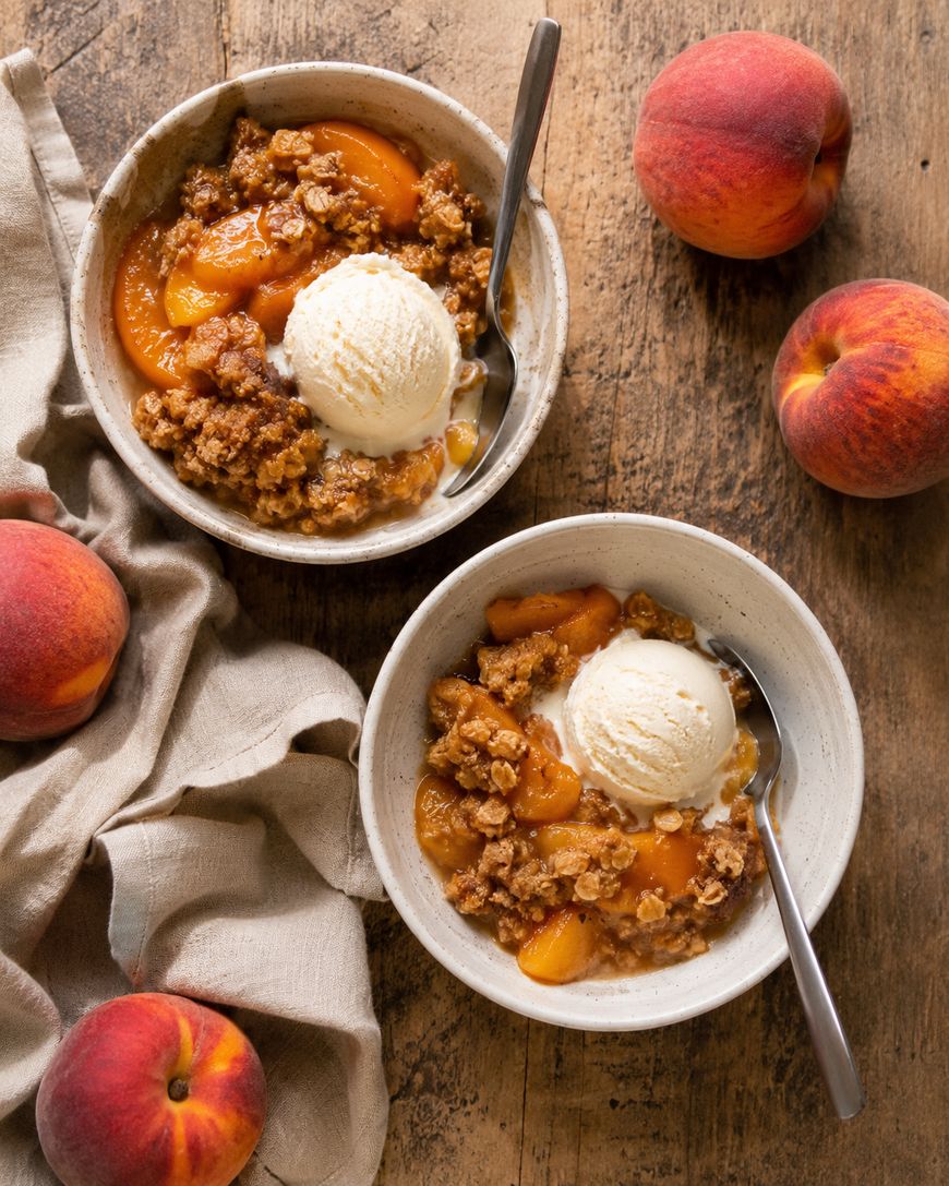 Two bowls of peach crumble topped with vanilla ice cream on a wooden table, surrounded by fresh peaches and a beige cloth. Spoons rest in each bowl.