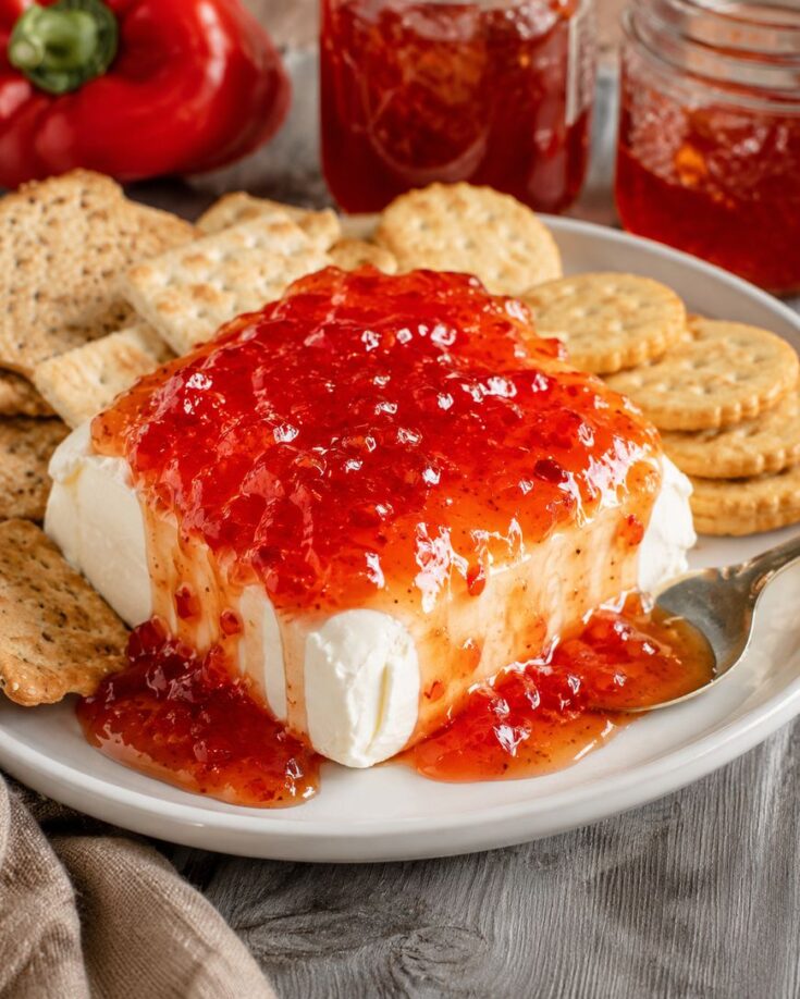 A block of cream cheese topped with red pepper jelly is surrounded by round crackers on a plate, with a spoon and jars of jelly nearby.