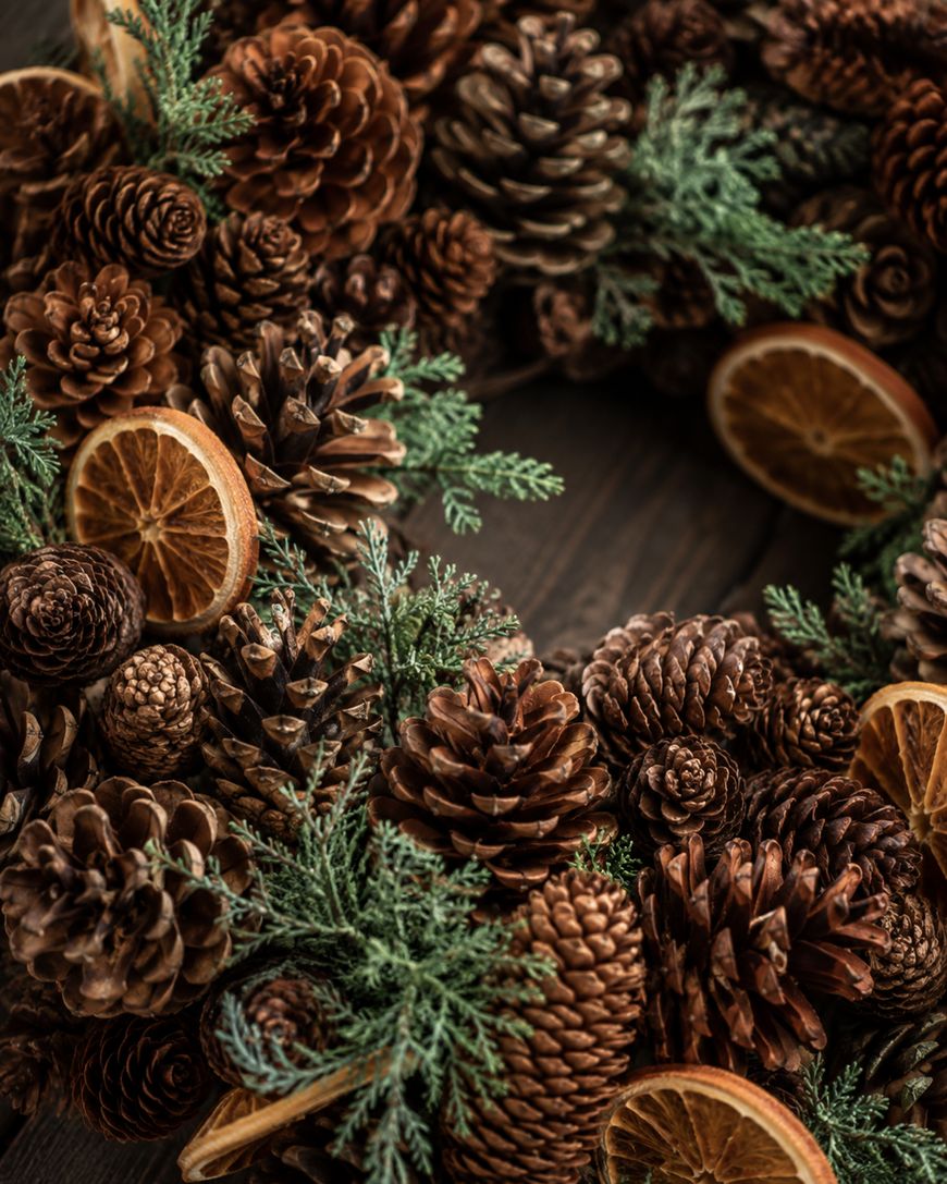 A festive wreath made of pine cones, dried orange slices, and green foliage sits on a dark wooden surface.