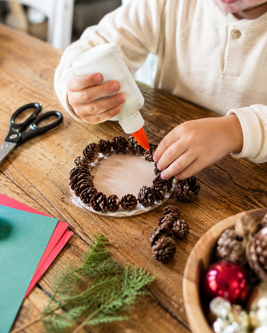 A child glues small pinecones onto a circular base for a craft project. Scissors, paper, evergreen sprigs, and ornaments are on the table.