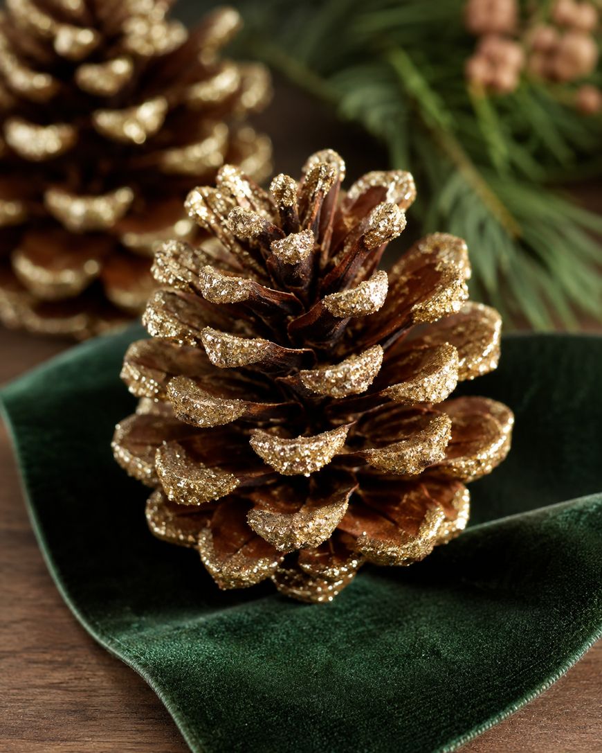 A pine cone with gold glitter on the tips sits on a green velvet ribbon, with pine needles and another pine cone blurred in the background.