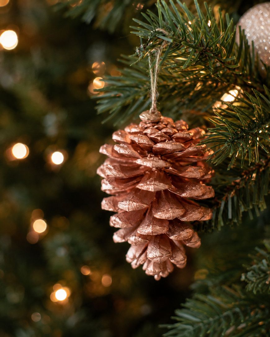 A painted pine cone ornament hangs from a green Christmas tree branch, with warm white lights softly glowing in the blurred background.