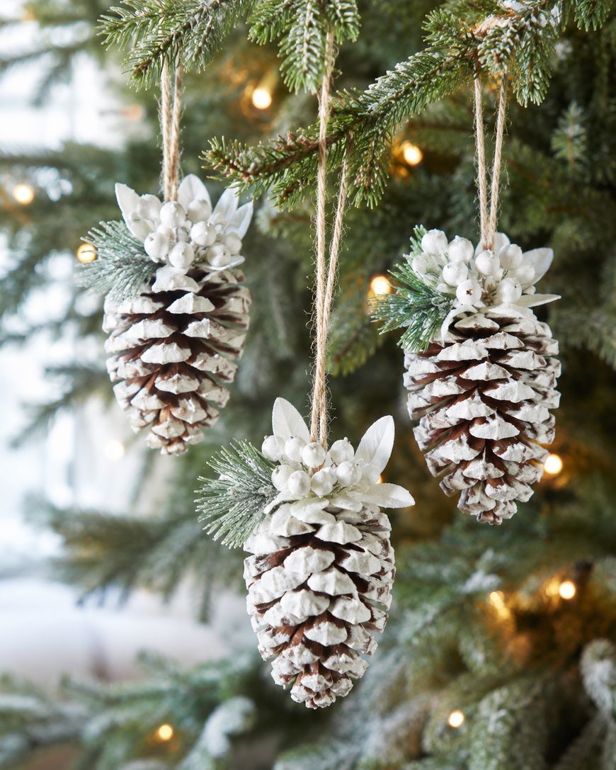 Three pine cone ornaments dusted with white, decorated with faux greenery and berries, hang from twine on a Christmas tree with glowing string lights in the background.
