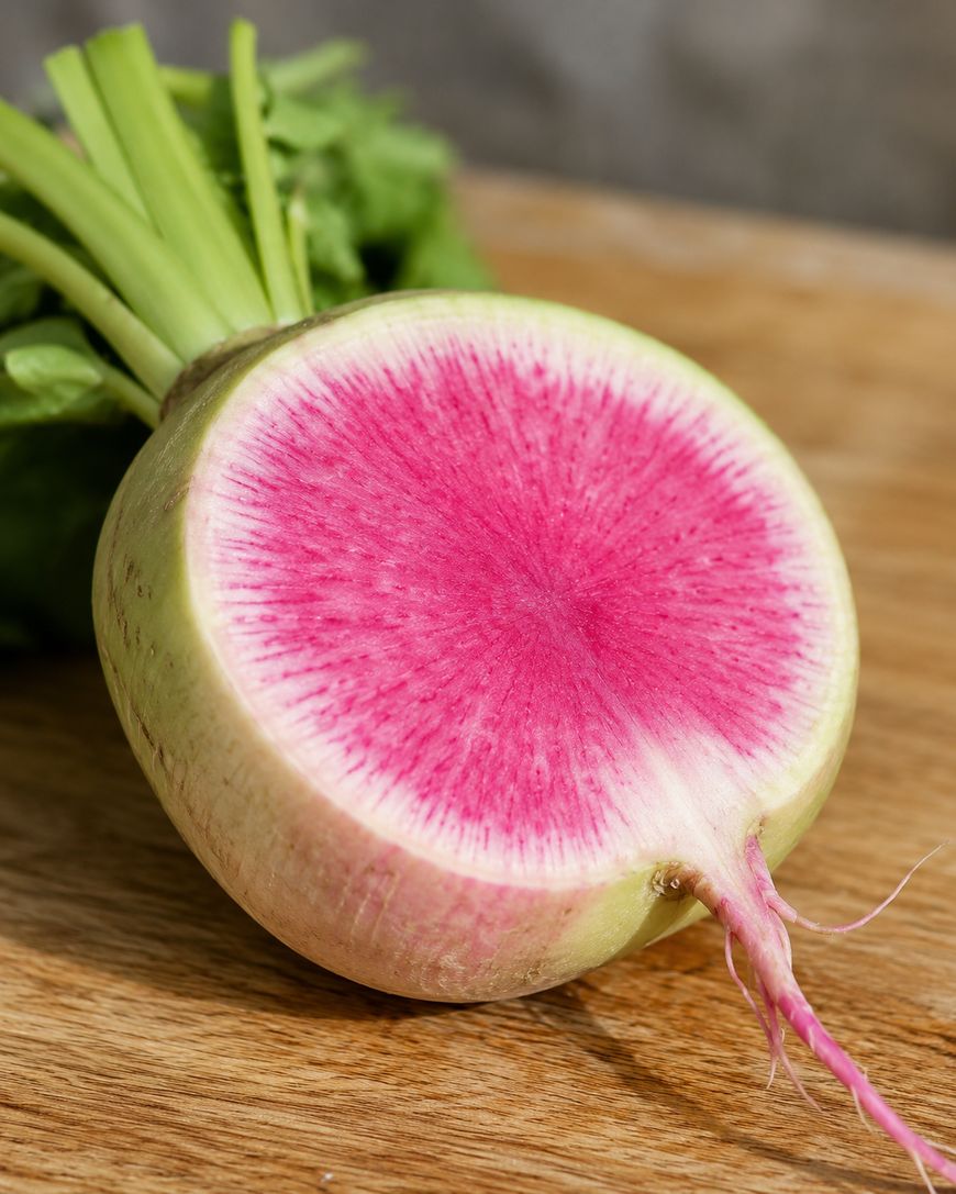 A sliced watermelon radish with a pale green outer skin and vibrant pink interior sits on a wooden surface, showing its colorful pattern and green leafy tops.