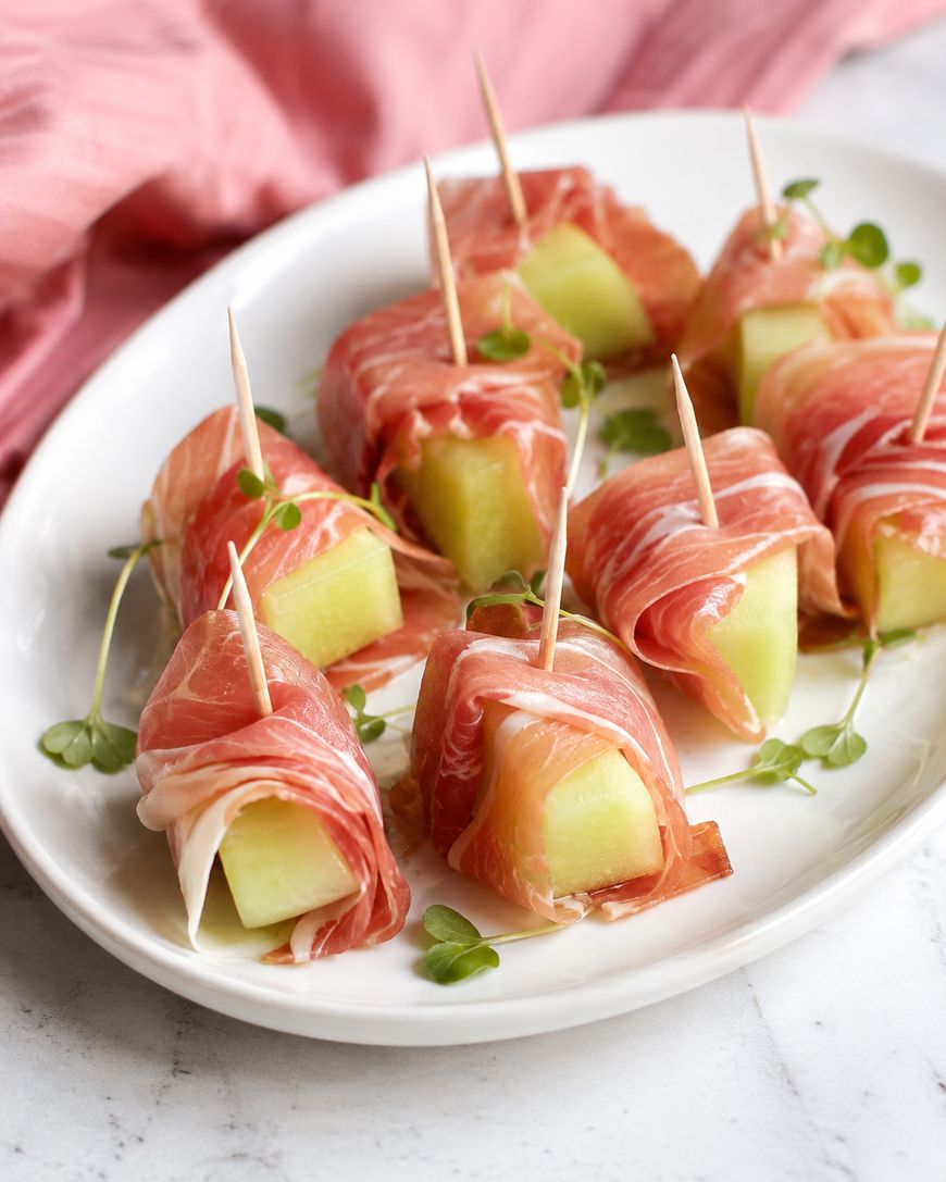 Eight pieces of prosciutto-wrapped melon are arranged on a white plate, each secured with a toothpick and garnished with microgreens, with a pink cloth in the background.