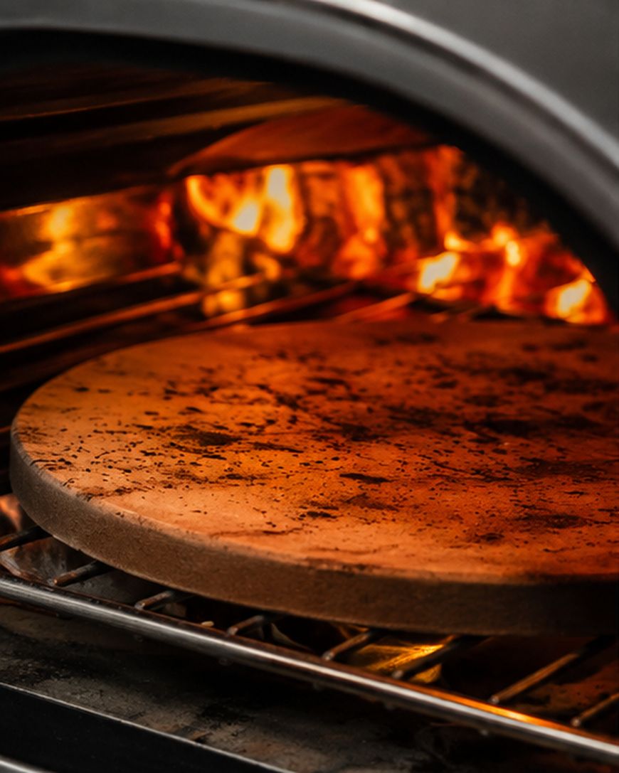 A round pizza stone is heating up inside an oven with visible flames in the background, ready for baking.