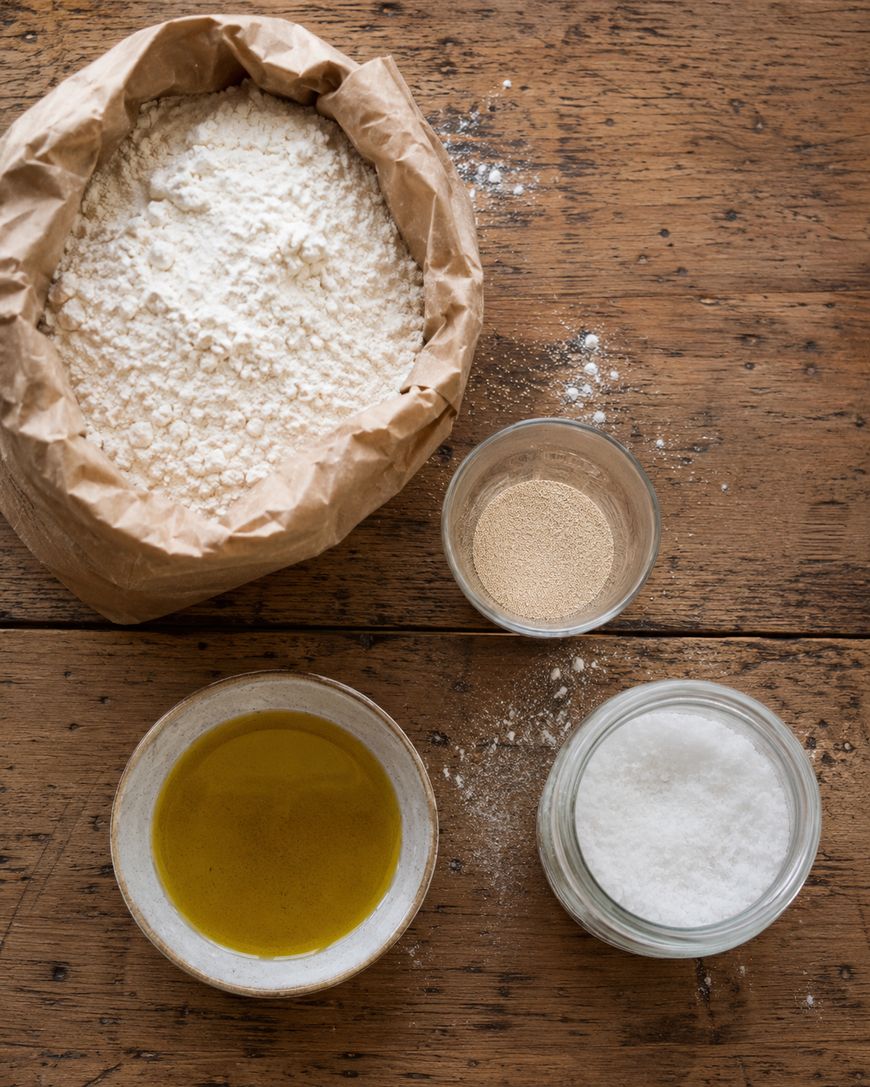 A brown paper bag of flour, a small bowl of olive oil, a glass jar of salt, and a small bowl of yeast are arranged on a rustic wooden table.