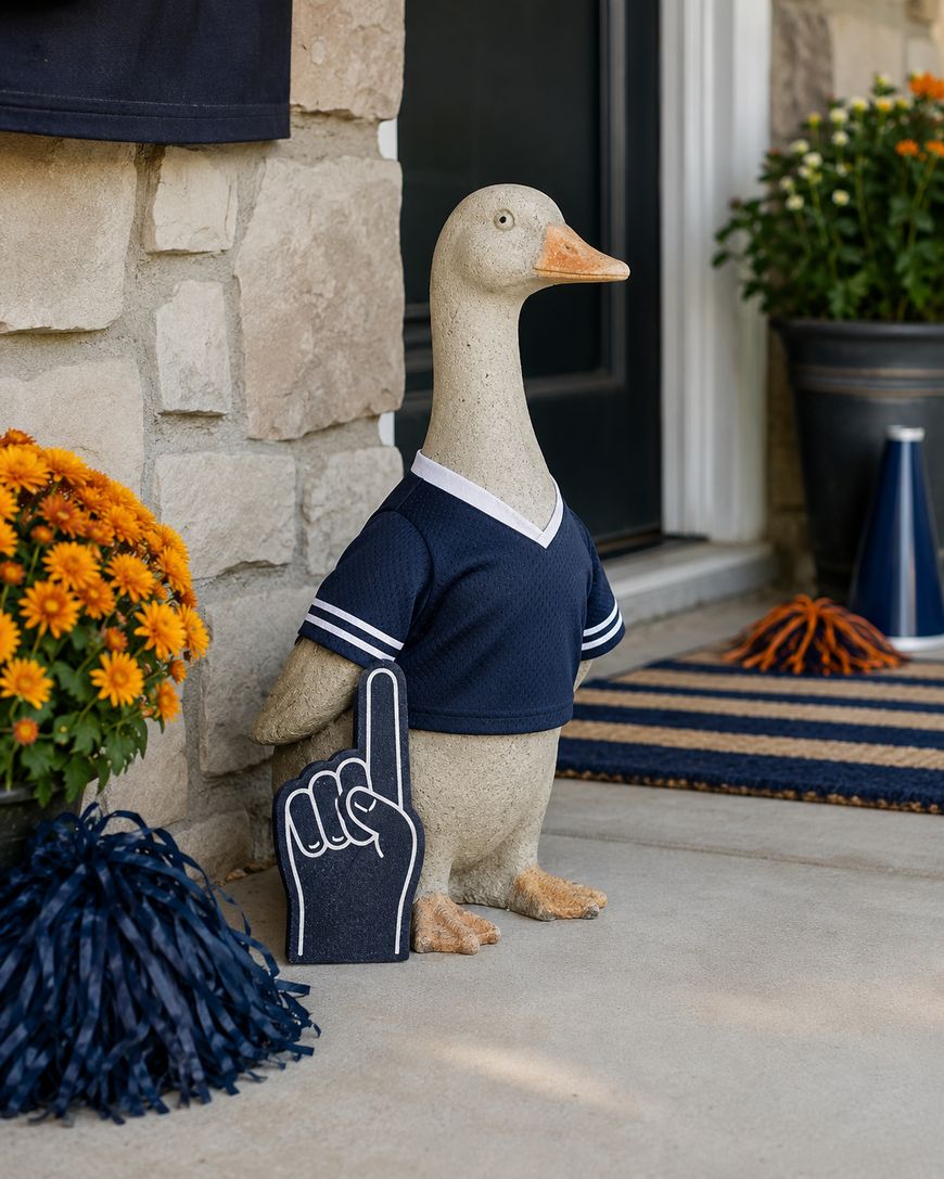 A goose statue dressed in a navy sports jersey stands on a porch, holding a foam finger. Orange flowers, a blue pom-pom, and a megaphone are nearby, creating a festive, sporty atmosphere.