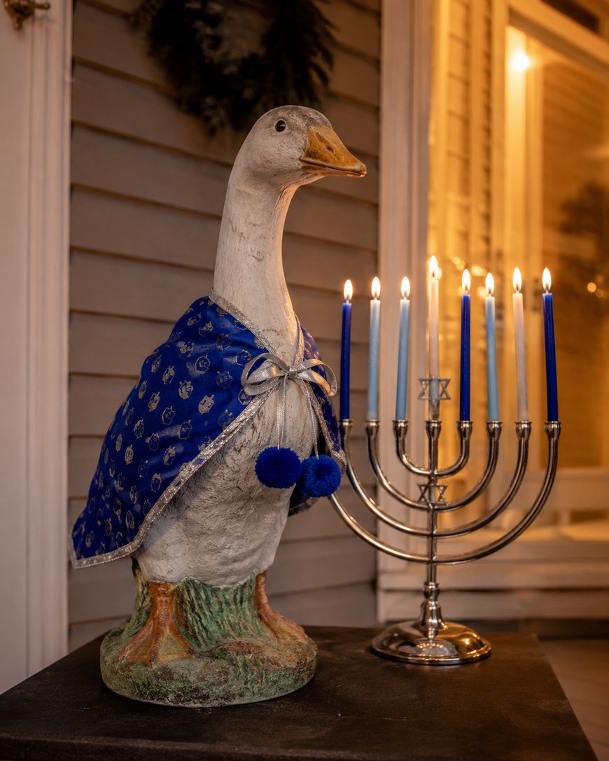 A statue of a goose wearing a blue cape sits beside a lit Hanukkah menorah with blue and white candles, in front of a house with warm light glowing from the window.