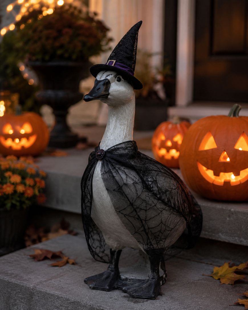 A goose statue dressed in a black witch hat and cape stands on a porch, surrounded by carved, glowing jack-o-lanterns and autumn leaves.