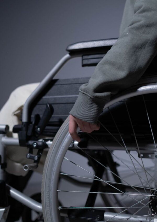 A person wearing a gray sweater rests their hand on the wheel of a wheelchair, positioned against a neutral background.