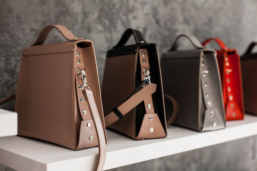 Several stylish handbags in beige, black, gray, and red are neatly lined up on a white shelf against a textured gray wall. Each bag features a side strap and metallic stud accents.