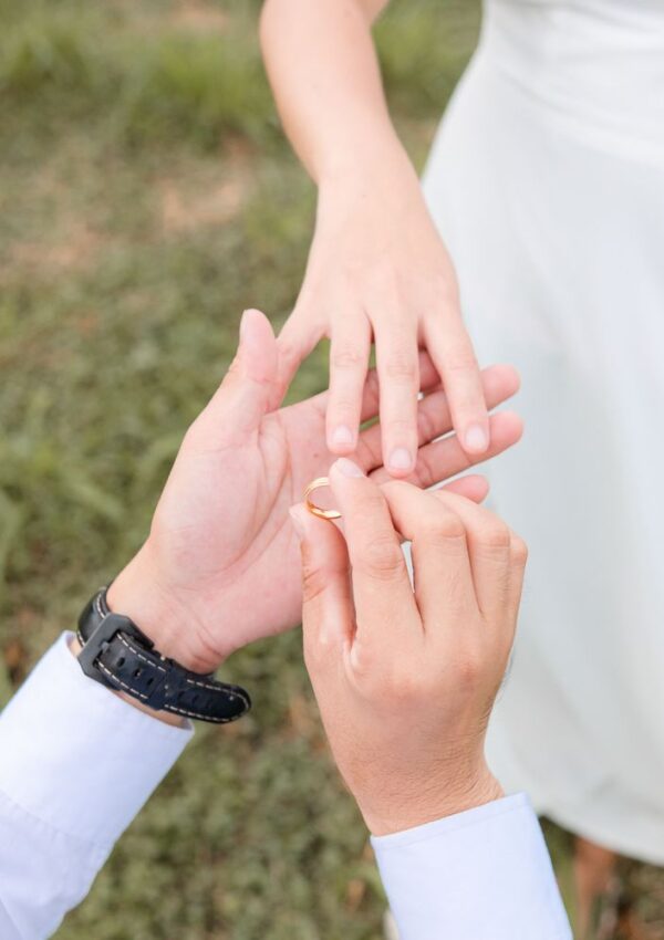 A person places a ring on another persons finger outdoors, suggesting a proposal or engagement; both wear white clothing.