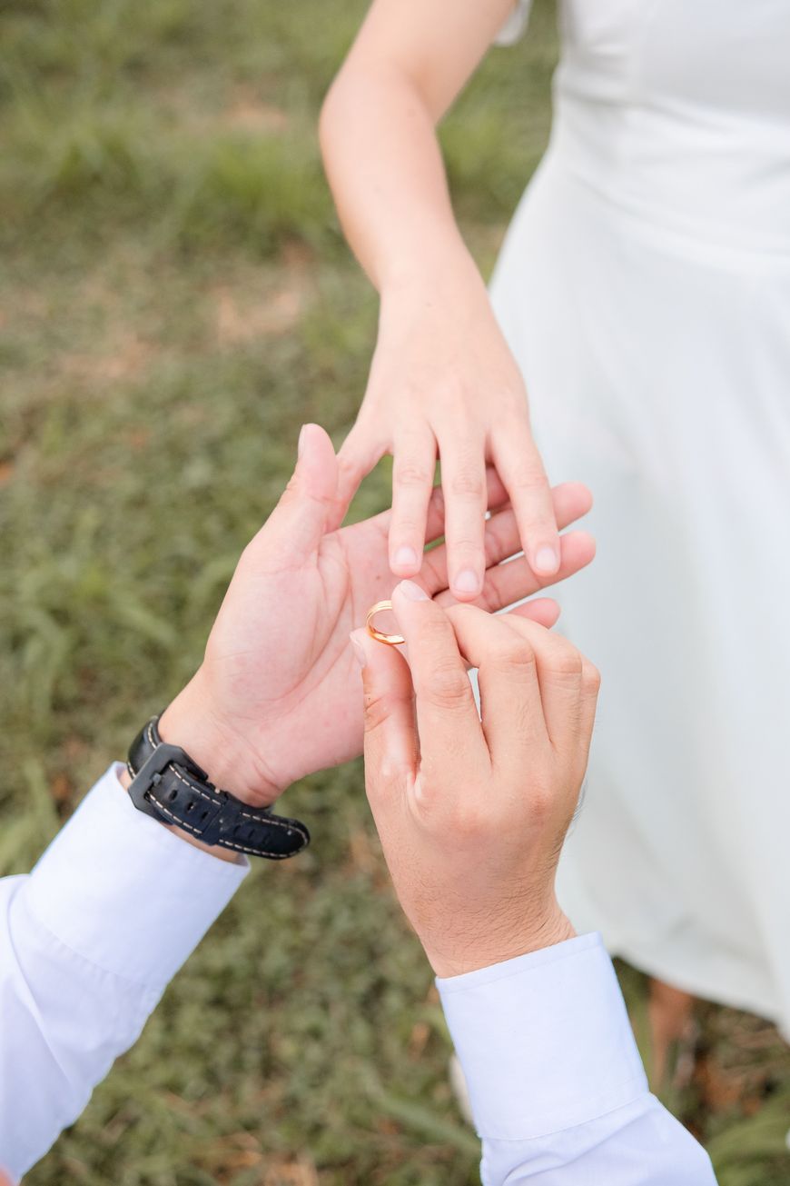 A person in a white shirt is placing a gold ring on another persons finger, who is wearing a white dress. They are outdoors on green grass.