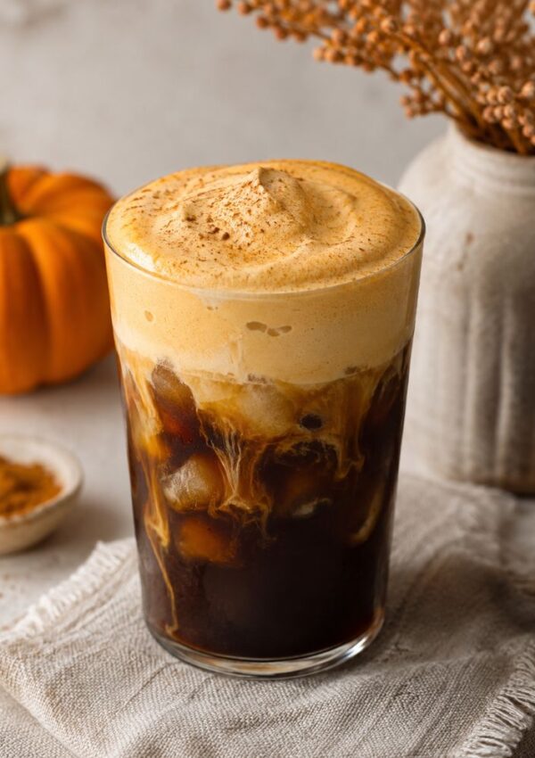 A glass of iced coffee with creamy foam on top, sprinkled with cinnamon, sits on a textured cloth with a small pumpkin and vase in the background.