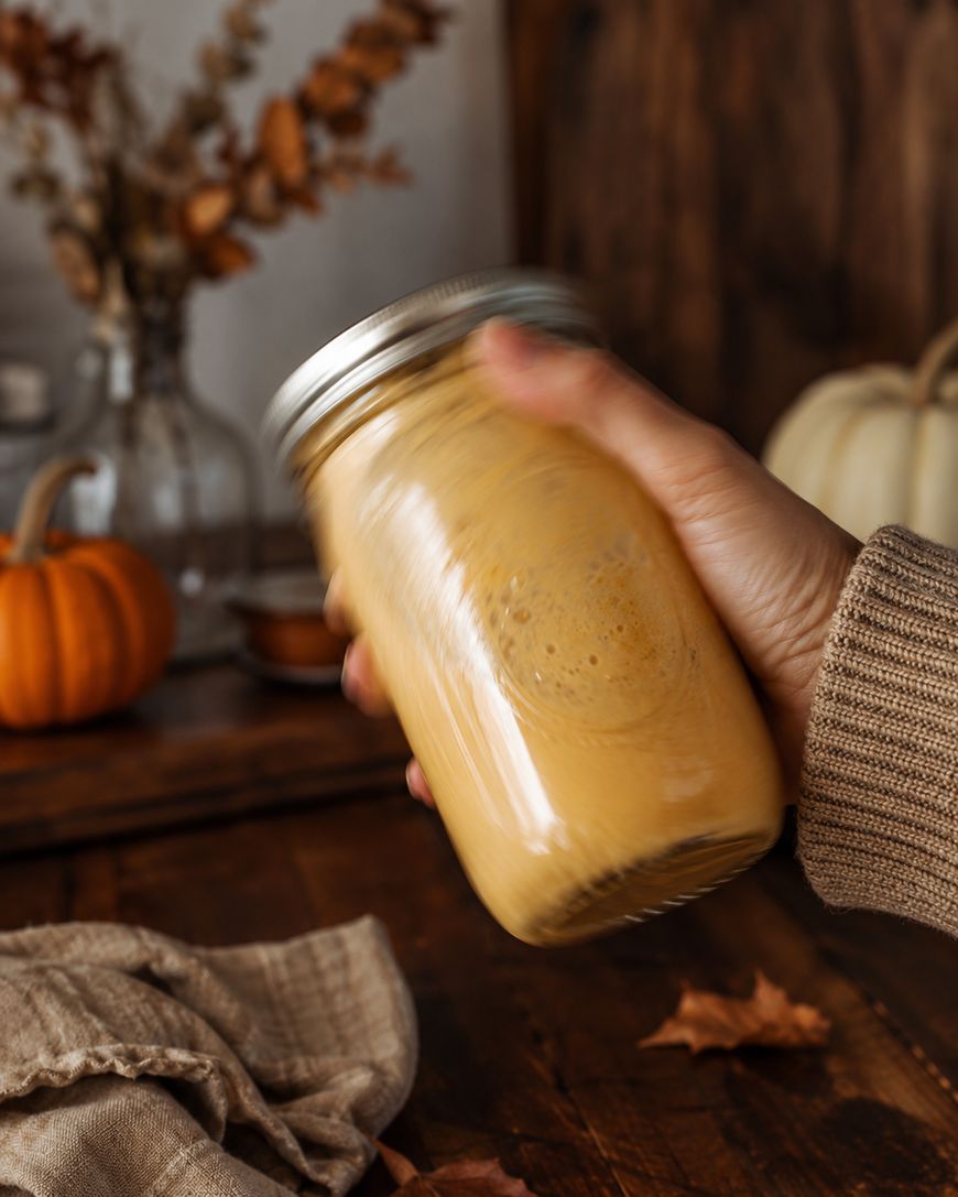 A hand in a brown sweater holds and shakes a mason jar filled with a creamy orange drink. There are autumn decorations, including pumpkins and dried leaves, on a wooden surface in the background.