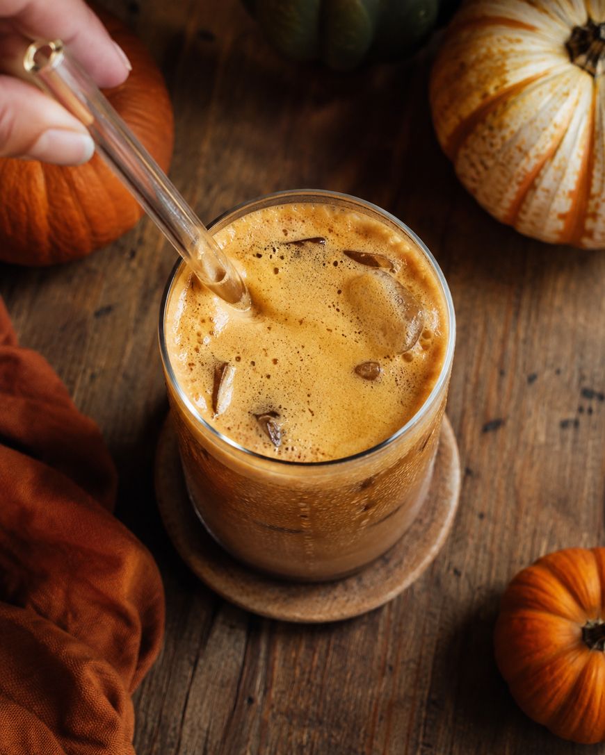 A glass of iced coffee with frothy foam on top sits on a wooden coaster, surrounded by small pumpkins on a rustic wooden table. A hand stirs the drink with a glass straw.