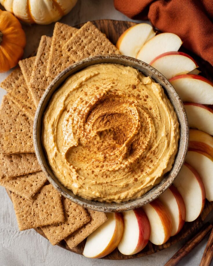 A bowl of creamy pumpkin dip, sprinkled with cinnamon, sits on a platter surrounded by graham crackers and sliced red apples. Small pumpkins and a brown napkin are nearby for decoration.
