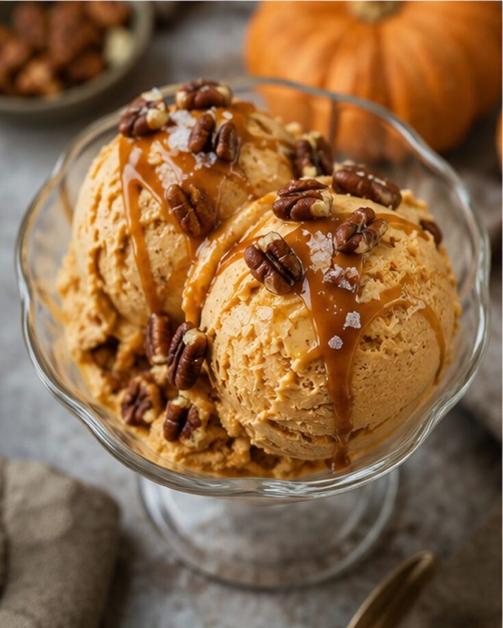 Two scoops of pumpkin ice cream in a glass dish, topped with pecans, caramel sauce, and sea salt, with a pumpkin and bowl of pecans blurred in the background.