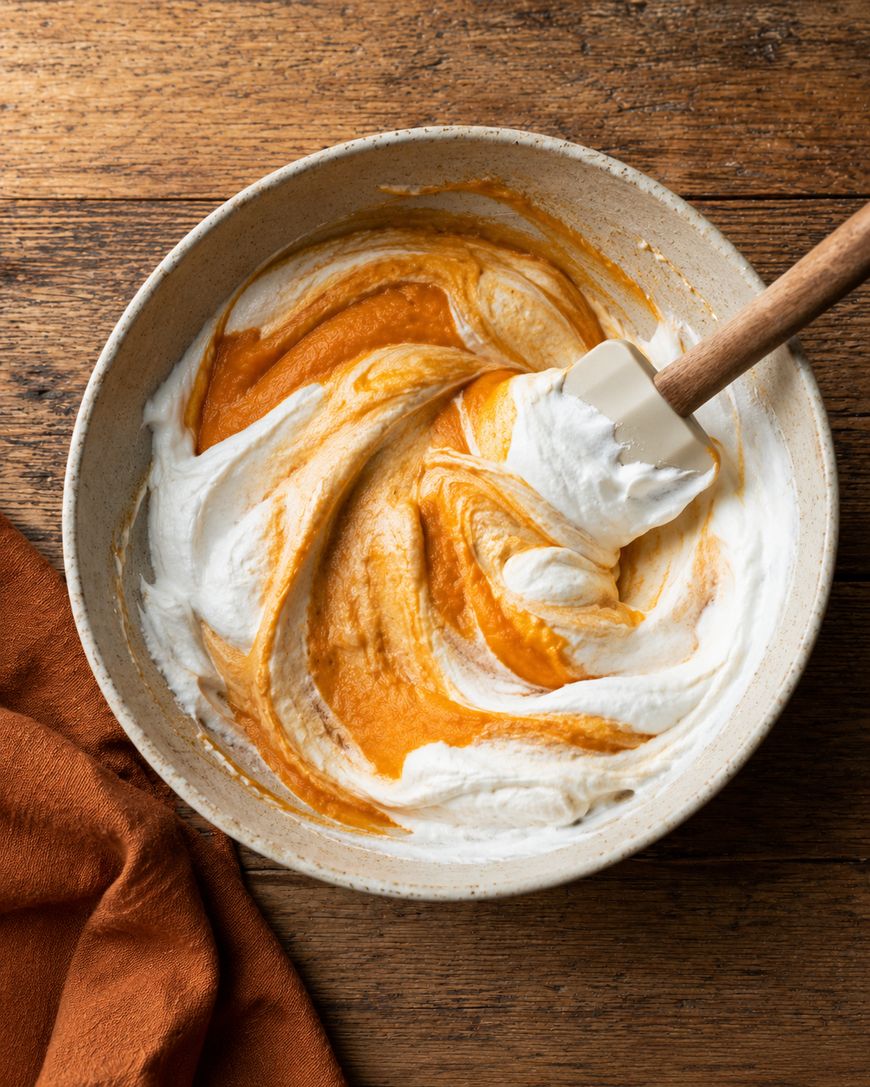 A mixing bowl with white whipped cream and orange pumpkin puree being folded together with a spatula on a wooden surface. An orange cloth is partially visible beside the bowl.