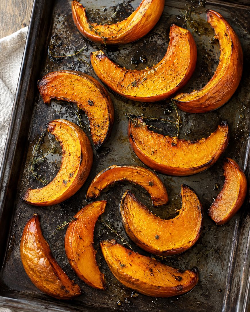 Roasted pumpkin wedges with herbs on a dark baking sheet, showing golden-brown, caramelized edges and scattered seasoning.