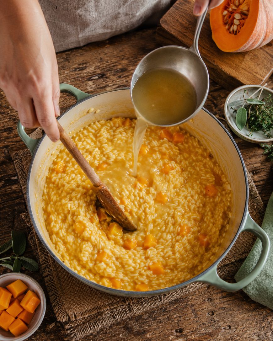 Person stirring creamy risotto with chunks of squash in a pot while pouring broth from a ladle. Fresh herbs, cubes of squash, and a sliced squash are on a rustic wooden table nearby.