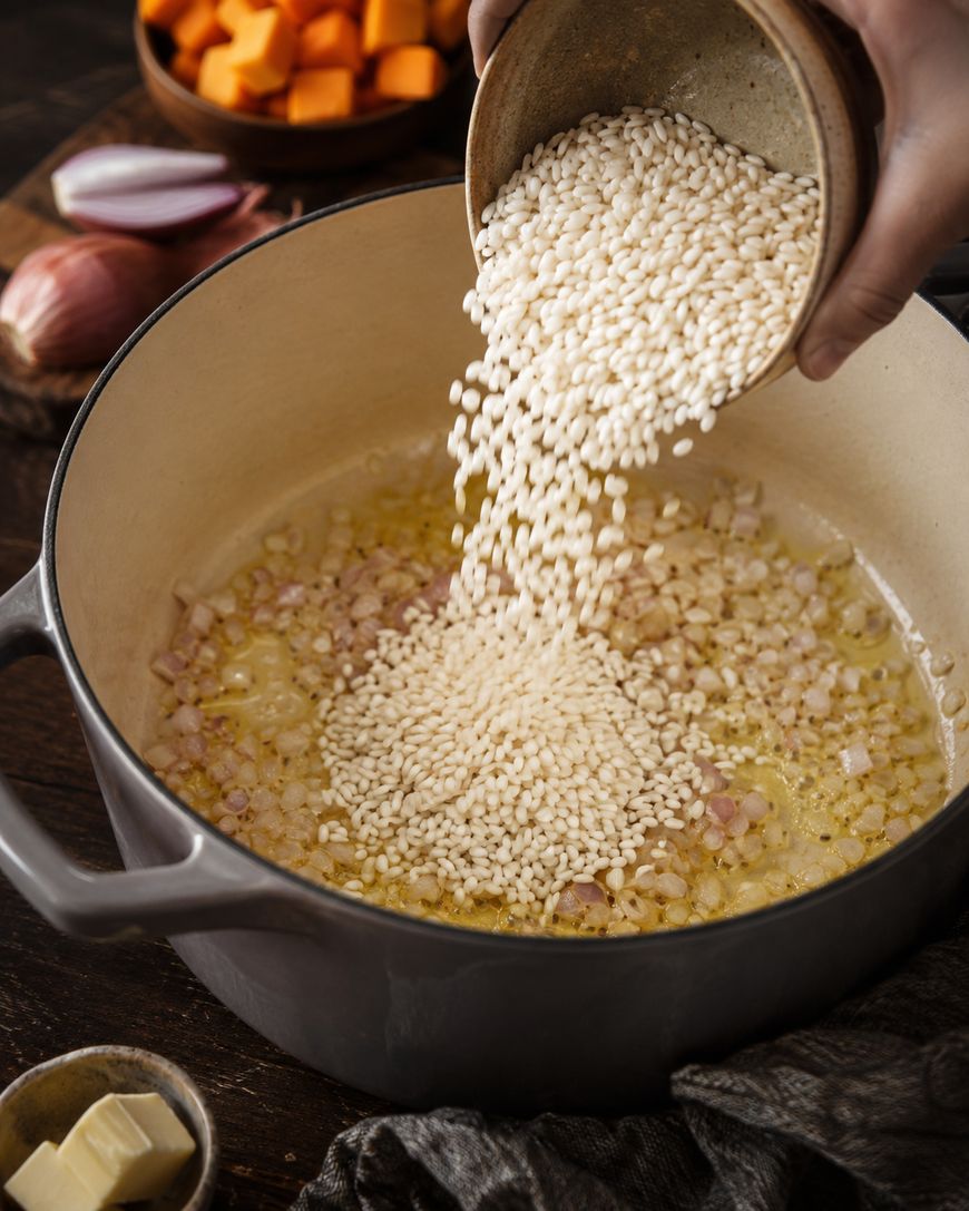 Rice is being poured into a pot with sautéed shallots and butter. In the background are diced butternut squash, a shallot, and a bowl of butter cubes on a wooden table.
