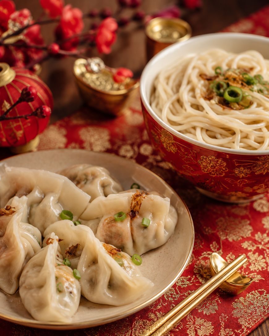 A plate of steamed dumplings garnished with green onions sits beside a bowl of noodles in broth, all arranged on a red and gold patterned cloth with festive decorations nearby.