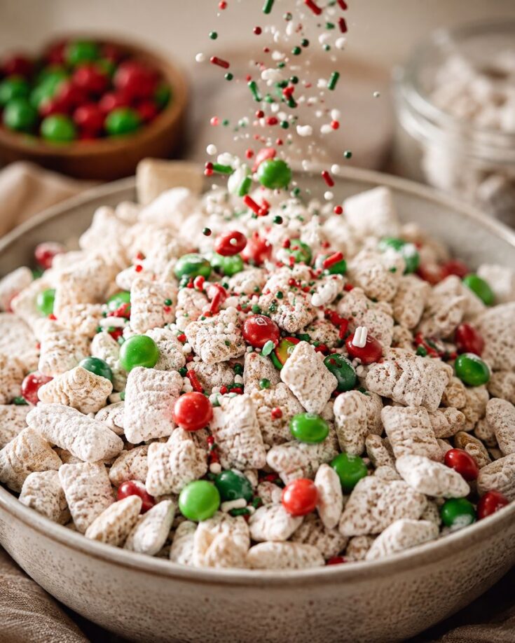 A bowl of powdered sugar-coated cereal mixed with red and green candies, with festive sprinkles falling on top. Blurred jars and bowls of candy are visible in the background.